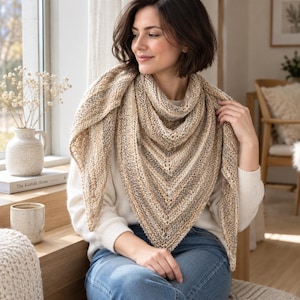 May include: A triangular knit shawl featuring a variegated color palette of beige, cream, brown, and grey tones. The accessory displays a textured stitch pattern with a subtle openwork design along the edges, draped over a white long-sleeve sweater.