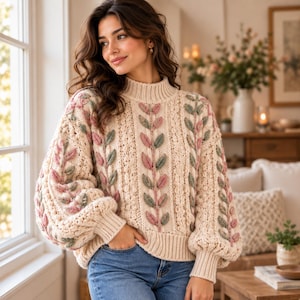 May include: A cream-coloured knit jumper featuring a high neck, ribbed cuffs, and a textured pattern of vertical vines with raised pink and sage green leaves. The garment has long, voluminous sleeves and is paired with blue denim jeans.