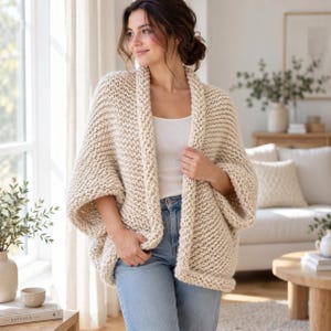Può includere: Un cardigan in maglia grossa color crema con apertura frontale, maniche larghe e una spessa trama a legaccio. Il capo è indossato sopra una semplice canotta bianca e jeans azzurri, presentando una silhouette ampia e rilassata.