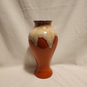 May include: A ceramic vase with a flared rim and a tapered base. The body features a glossy, solid burnt orange finish on the lower portion, transitioning to a textured, cream-colored drip glaze that cascades from the rim down the neck.