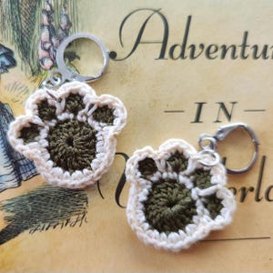 May include: Two crocheted paw print keychains with a white border and a dark green center. The keychains are attached to silver key rings.