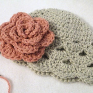 May include: A handmade crochet hat in a light beige colour, adorned with a large, peach-coloured crochet flower. The flower is intricately designed with multiple layers of petals. A needle and yarn are visible, suggesting the item is in progress.
