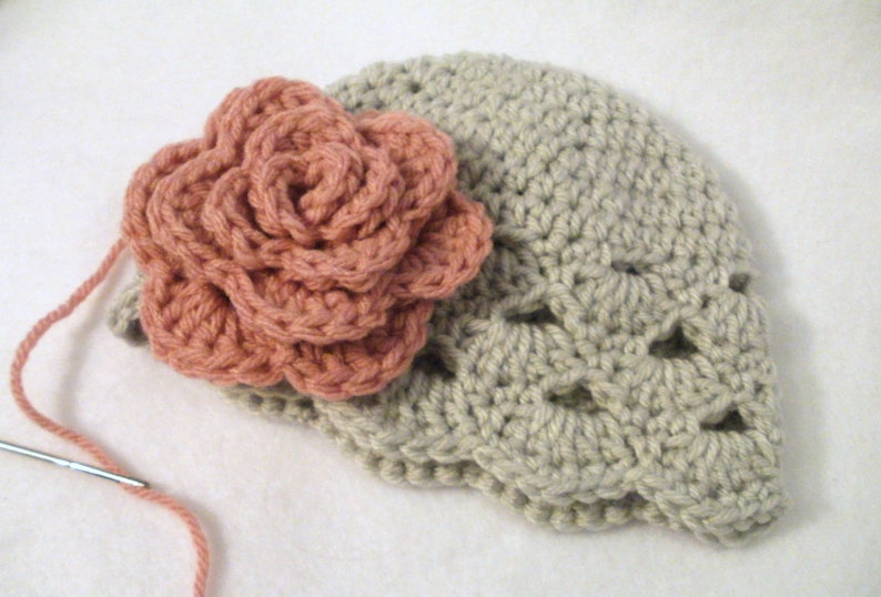 May include: A handmade crochet hat in a light beige colour, adorned with a large, peach-coloured crochet flower. The flower is intricately designed with multiple layers of petals. A needle and yarn are visible, suggesting the item is in progress.