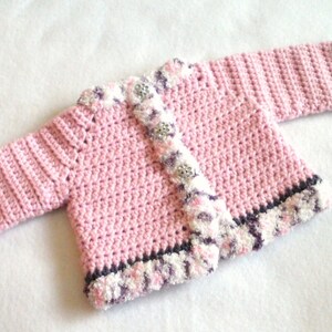 May include: A pink crocheted baby cardigan with a white and purple trim. The cardigan has a button closure and a textured stitch pattern.