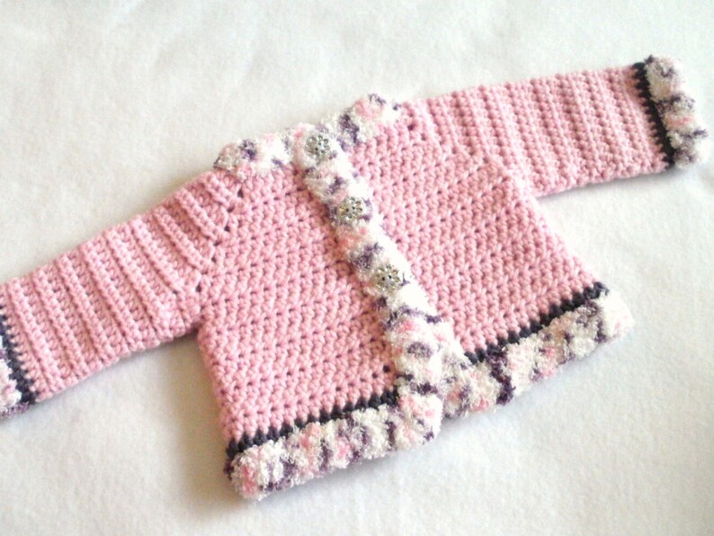 May include: A pink crocheted baby cardigan with a white and purple trim. The cardigan has a button closure and a textured stitch pattern.