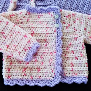 May include: A crocheted baby cardigan with a pink and white color scheme. The cardigan has a scalloped edge and a single button closure.
