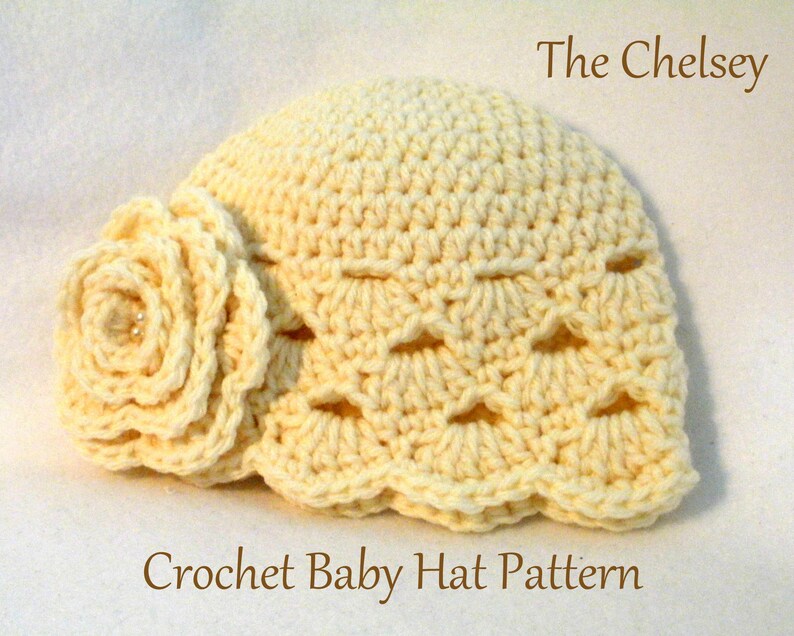 May include: A cream-coloured crocheted baby hat with a decorative flower and scalloped edge. The hat has an openwork design and is labelled "The Chelsey" and "Crochet Baby Hat Pattern". The hat is made of yarn.
