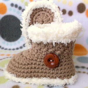 K&ouml;nnte beinhalten: Braune geh&auml;kelte Babyschuhe mit einem wei&szlig;en flauschigen Besatz und einem braunen Knopf. Die Schuhe liegen auf einem wei&szlig;en und braunen gemusterten Hintergrund.