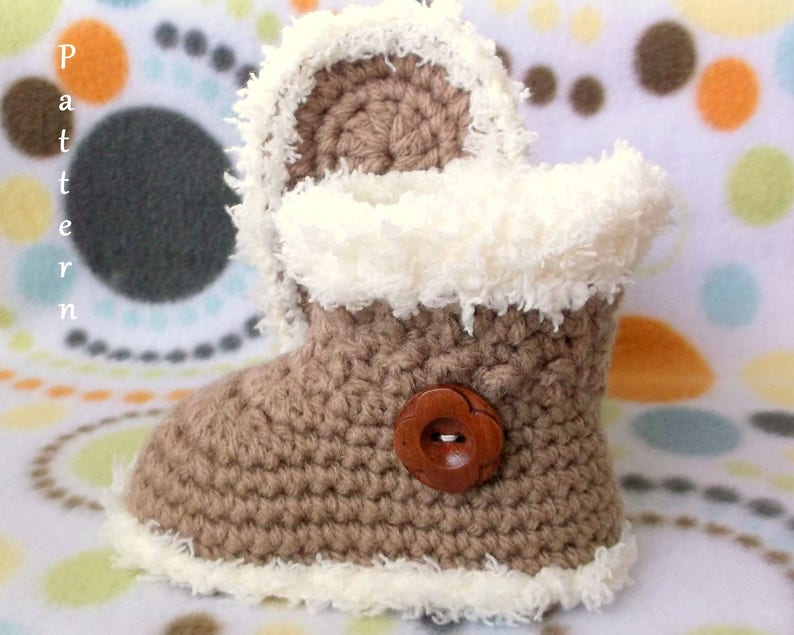 K&ouml;nnte beinhalten: Braune geh&auml;kelte Babyschuhe mit einem wei&szlig;en flauschigen Besatz und einem braunen Knopf. Die Schuhe liegen auf einem wei&szlig;en und braunen gemusterten Hintergrund.