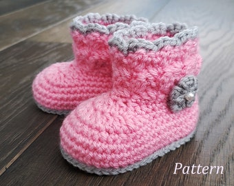 Puede incluir: Un par de botines de bebé de crochet rosa con ribete gris y una flor gris como adorno. Los botines están sobre una superficie de madera.