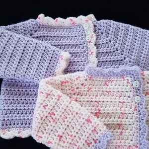May include: Two crocheted baby sweaters, one in lavender and one in white with pink accents. Both sweaters have a scalloped edge and button closures.