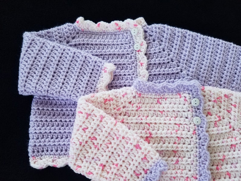May include: Two crocheted baby sweaters, one in lavender and one in white with pink accents. Both sweaters have a scalloped edge and button closures.
