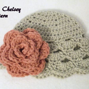 May include: A light grey crocheted hat with a peach-coloured crocheted flower. The hat has a scalloped edge and the text "The Chelsey Pattern" is visible in the upper left corner.
