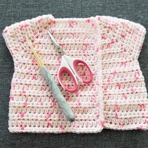 May include: A pink and white crocheted vest with a crochet hook and scissors lying on top of it.