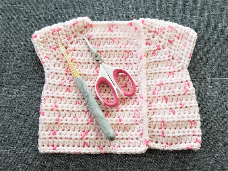 May include: A pink and white crocheted vest with a crochet hook and scissors lying on top of it.