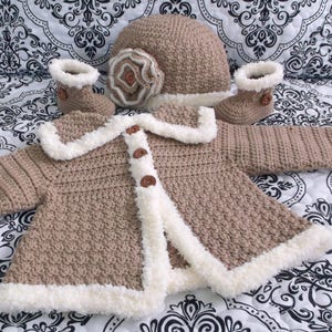 K&ouml;nnte beinhalten: Ein geh&auml;keltes Baby-Outfit in Braun und Wei&szlig;. Das Set beinhaltet eine M&uuml;tze, eine Jacke und Booties. Die Jacke hat einen wei&szlig;en Pelzbesatz und braune Kn&ouml;pfe. Die M&uuml;tze hat eine wei&szlig;e Blumenverzierung.