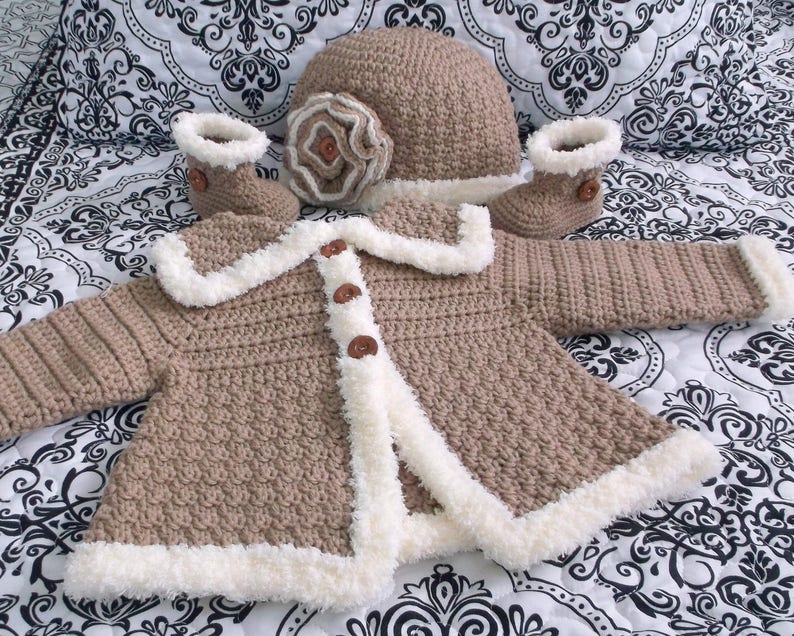 K&ouml;nnte beinhalten: Ein geh&auml;keltes Baby-Outfit in Braun und Wei&szlig;. Das Set beinhaltet eine M&uuml;tze, eine Jacke und Booties. Die Jacke hat einen wei&szlig;en Pelzbesatz und braune Kn&ouml;pfe. Die M&uuml;tze hat eine wei&szlig;e Blumenverzierung.