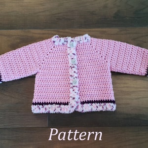 May include: Pink crocheted baby cardigan with a white and purple trim. The cardigan has a button closure and is made with a soft yarn.