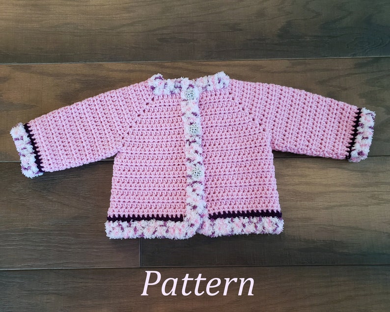 May include: Pink crocheted baby cardigan with a white and purple trim. The cardigan has a button closure and is made with a soft yarn.