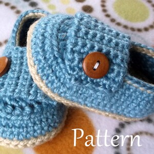 May include: A pair of blue crocheted baby booties with a beige trim and wooden buttons. The booties are on a white and brown patterned fabric.