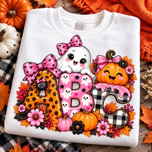Op de afbeelding: Een wit sweatshirt met een afbeelding van de letters A, B en C. De A is oranje met zwarte stippen, de B is roze met witte spookjes en de C is zwart-wit geblokt met roze glazuur. Bevat pompoenen, bloemen en een spookje.