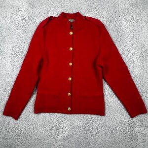 May include: A bright red long-sleeve knit cardigan featuring a mock neck collar, shoulder epaulets, and a front button closure with six round gold-toned metal buttons. The garment has a straight hemline and two small horizontal slit pockets.