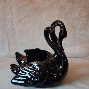 May include: A glossy black ceramic swan planter featuring a curved neck and a hollow body. The swan is depicted with its head arched gracefully, creating an open loop shape. The surface is smooth and reflective, showcasing a deep black finish.