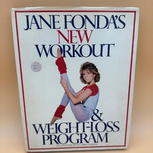 May include: A rectangular book titled Jane Fonda's New Workout & Weight-Loss Program with a white cover and red border. The cover features a photograph of a person in a grey leotard with red sleeves, light blue leggings, and red leg warmers.
