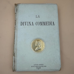 May include: A vintage book titled La Divina Commedia with a light gray cover. The front features black text and a circular gold-toned medallion depicting a profile portrait. At the bottom, the text reads Ulrico Hoepli Editore Libraio Della Real Casa Milano.