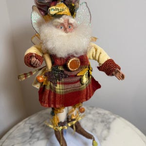 May include: A decorative figurine of a bearded character wearing a red and yellow plaid tunic, cream-colored sleeves, and brown pointed shoes with small bells. The figure features iridescent wings, a textured hat, and autumn-themed embellishments.