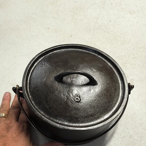 May include: A round, dark-colored cast iron pot with a matching lid. The lid features a central arched handle and the embossed number 8. A metal bail handle is attached to the sides of the pot, allowing for easy lifting and carrying of the cookware.