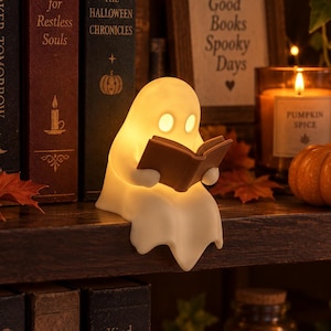 May include: A small, white, glowing ghost figurine sitting on a shelf edge, holding and reading a brown open book. The ghost has two oval-shaped eye cutouts and emits a warm, soft light from within its smooth, rounded, spectral body shape.