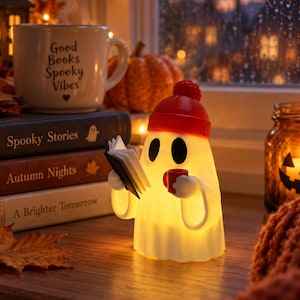 May include: A glowing white ghost figurine wearing a red beanie, holding a small open book and a red mug. The ghost has large black oval eyes and is illuminated from within, casting a warm light on the surface it rests upon.