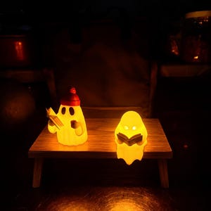 May include: Two glowing, translucent yellow ghost figurines positioned on a small wooden table. The left ghost wears a red beanie, holds a book, and a small mug. The right ghost is shorter, rounded, and holds an open book. Both emit a warm light.