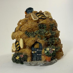 May include: A decorative beehive-shaped structure with a textured straw-like exterior, featuring a dark blue arched door with a bee emblem, green ivy vines, small colorful flowers in a pot, and several circular windows with yellow and black stripes.