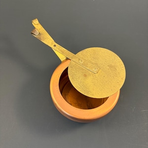 May include: A small, round copper container with a polished brass lid attached to a long, thin brass handle. The lid is circular and sits partially open, revealing the hollow interior of the copper vessel. The metal surfaces show a warm, metallic finish.
