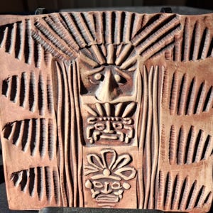 May include: A square terracotta-colored clay tile featuring a relief carving of a central figure with a sunburst headdress above two smaller stacked faces. The surface is decorated with vertical textured lines radiating outward from the center.