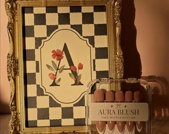 May include: A gold ornate picture frame containing a black and white checkered print with a large letter A and floral illustrations. Next to it is a clear plastic case of press-on nails labeled Aura Blush Nails, Beauty & Self-Care.