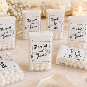 Peut inclure: Récipients rectangulaires transparents en plastique remplis de menthes blanches. Chaque étiquette blanche porte le texte noir Maxine and Juno ou Mint to Be, avec un dessin au trait de deux mains tenant une petite bague.