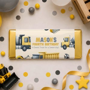 May include: A rectangular chocolate bar wrapped in gold foil with a cream-colored paper label featuring yellow construction trucks, a wrench, and the text Mason's Fourth Birthday A Sweet Treat for a Sweet day printed in gray and yellow.