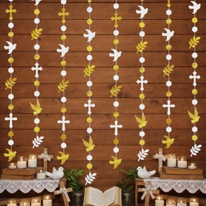 May include: A vertical garland set featuring alternating white and gold paper cutouts of crosses, doves, and leafy branches strung together. The decorative hanging strings are arranged in multiple rows against a dark brown wooden background.