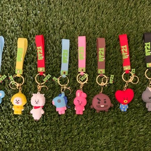 May include: A set of eight BT21 character keychains, each featuring a silicone wrist strap in various colors including light blue, yellow, red, pink, brown, and black, attached to a gold-tone metal ring with a small hanging character figure.