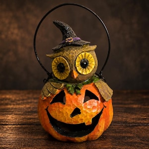 Puede incluir: Una figura decorativa que muestra un búho marrón con un sombrero de bruja negro con una banda morada, posado sobre una calabaza naranja tallada con una cara ancha y sonriente. La pieza incluye un asa de metal negro arqueada sobre el búho.