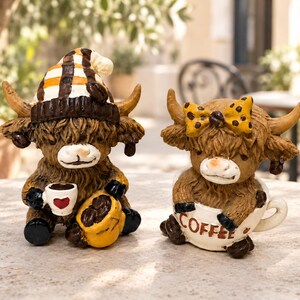 Puede incluir: Dos figuras de vacas highland marrones. La vaca de la izquierda lleva un gorro de cuadros marrón y blanco, sostiene una pequeña taza blanca con un corazón rojo y se sienta junto a un saco amarillo de granos de café. La derecha está en una taza COFFEE.