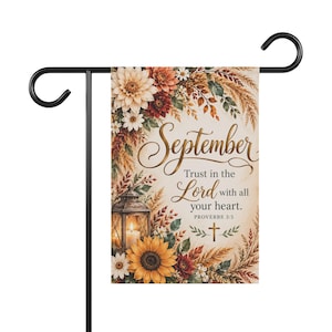 Könnte beinhalten: Eine rechteckige Gartenflagge an einem schwarzen Metallständer mit herbstlichem Blumenmotiv, Sonnenblumen, Dahlien und einer Laterne. Der Text lautet: September, Trust in the Lord with all your heart. PROVERBS 3:5, ergänzt durch ein kleines Kreuz.