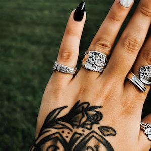 May include: A collection of silver-toned metal rings crafted from vintage silverware, featuring intricate floral embossed patterns and adjustable bands, worn on fingers with black and white nail polish and a black ink hand tattoo.