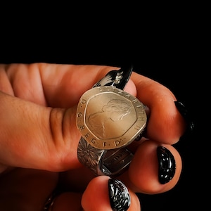 May include: A silver-toned ring crafted from a British coin featuring the profile of Queen Elizabeth II. The coin is inscribed with ELIZABETH II D G REG F D. The band of the ring displays a detailed floral pattern embossed into the metal surface.