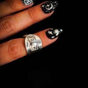 May include: A silver-toned metal ring featuring a raised cat figure on the band. The ring is worn on a finger with black almond-shaped nails decorated with white painted designs including a spider web, a crescent moon, stars, and bats.