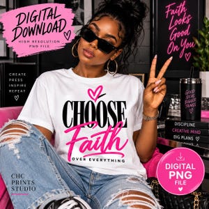 May include: A white t-shirt featuring the black text CHOOSE and the pink script text Faith, with the words OVER EVERYTHING in smaller black text below. A pink heart icon is positioned above the word CHOOSE. The design is centered on the chest.