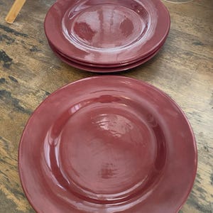 May include: A set of three round dinner plates with a glossy, deep maroon finish. The plates feature a smooth, slightly concave center and a wide, flat rim. The surface of the plates has a subtle, organic texture with visible variations in the glaze.